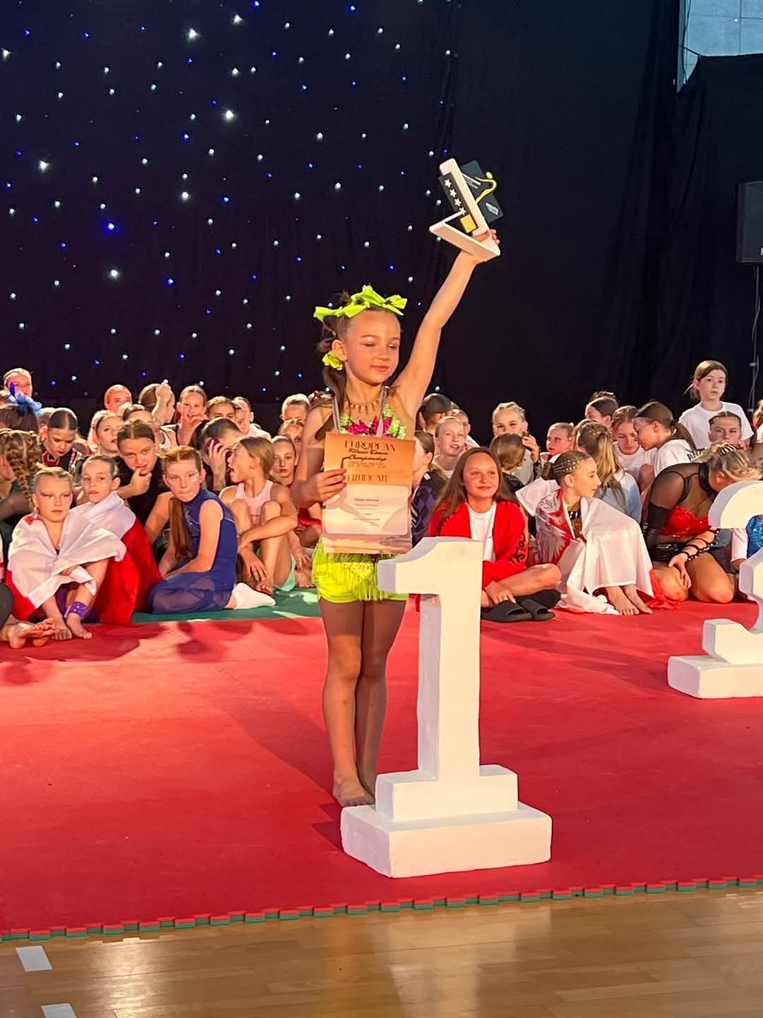 Mała dziewczynka w neonowym, żółto-różowym stroju sportowym stoi na najwyższym stopniu podium z numerem 1. W jednej ręce trzyma dyplom, a drugą wznosi do góry, trzymając statuetkę. W tle siedzi grupa innych dzieci na czerwonym dywanie.